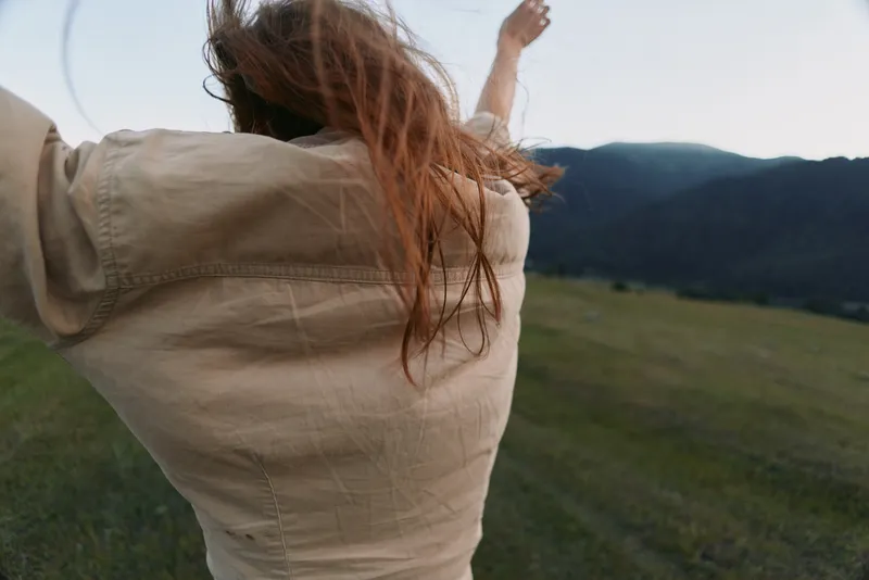 Woman outdoors in a wide landscape field beside mountains, arms raised, back view, conveying joy, freedom and a relaxed lifestyle in natural light