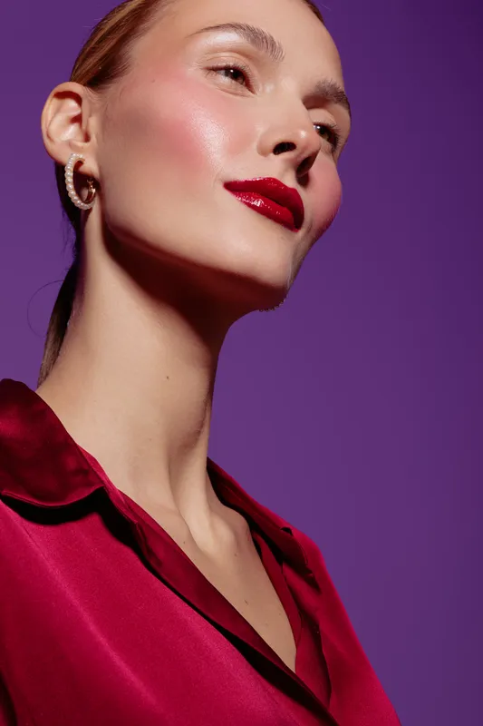 Close-up of a confident woman with glossy red lips and smooth skin wearing a red satin blouse against a purple background. Concept of beauty, makeup, and fashion portrait.
