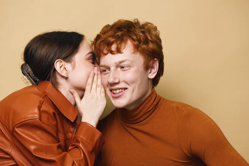 Young woman whispering secret to smiling redhead man in cozy autumn clothing on beige background. Friendly communication and happy expression in casual studio portrait.