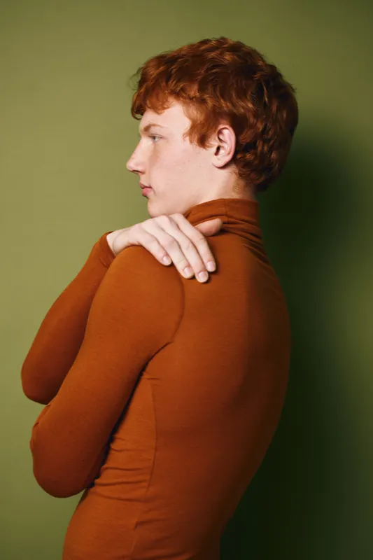 Young man with red hair in brown turtleneck sweater posing sideways against green background with hands touching shoulder in contemplative mood.