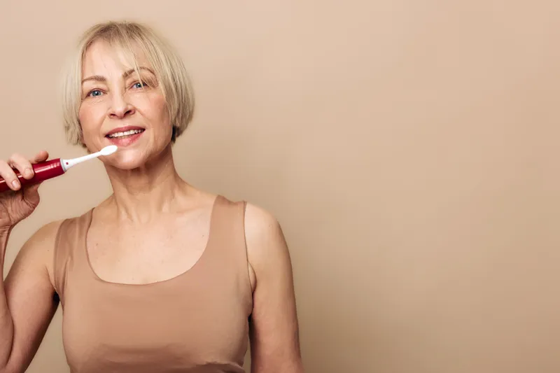 Mature woman with short blonde hair brushes teeth with electric toothbrush in beige tank top. Clean skincare and dental hygiene concept with natural light and warm neutral background.