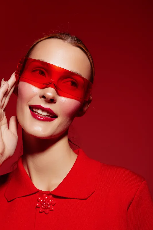 Smiling woman in red sweater and red translucent glasses poses on red background. Fashion portrait with bright makeup and stylish accessories. Concept of modern style and confidence.