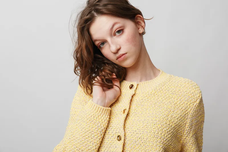 Young woman with curly hair wearing yellow knitted sweater looks thoughtfully at camera on light background. Concept of casual fashion, natural beauty, and soft texture clothing.