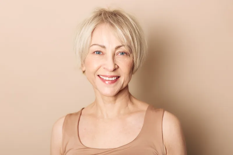 Smiling mature woman with short blonde hair wears beige top against neutral background. Concept of confidence, natural beauty, aging gracefully, and positive lifestyle for seniors.