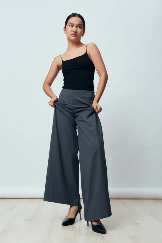 Woman in black tank top and wide leg gray pants standing with hands in pockets against plain background looking sideways confident stylish fashion model indoor.