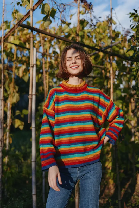 Woman in her 20s smiling, wearing a colorful striped sweater and casual outfit, young adult standing in a sunlit garden by trellis, bob haircut, relaxed lifestyle and fashion portrait.