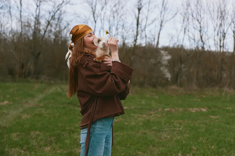 A woman outdoors in a brown jacket and orange beanie kisses a goat kid in a grassy field, capturing affection and bond with animals, a simple lifestyle in nature, warm light, rustic charm, and a