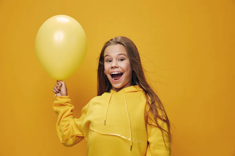 Happy girl in yellow hoodie holding yellow balloon for celebration, birthday or party event with excitement and joy on yellow background in studio.