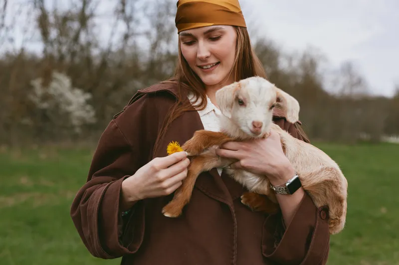 A woman in a brown coat and headscarf holds a small goat kid outdoors, admiring a flower. The scene conveys gentle interaction, rural lifestyle, and compassion for animals.