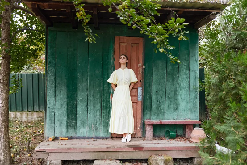 woman standing on cottage porch in rural setting vintage dress wooden green facade nature authenticity inclusivity