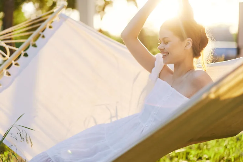 Young woman enjoying a sunny day in a hammock, wearing a white summer dress, radiating happiness in a vibrant green outdoor setting with bright sunlight