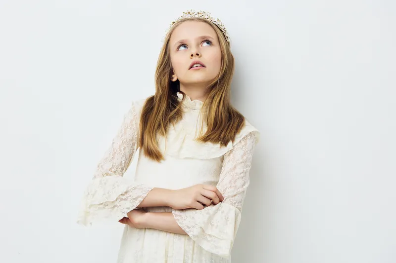 Young girl in a white dress and crown, looking thoughtfully upwards against a light background, capturing innocence and curiosity in a serene moment