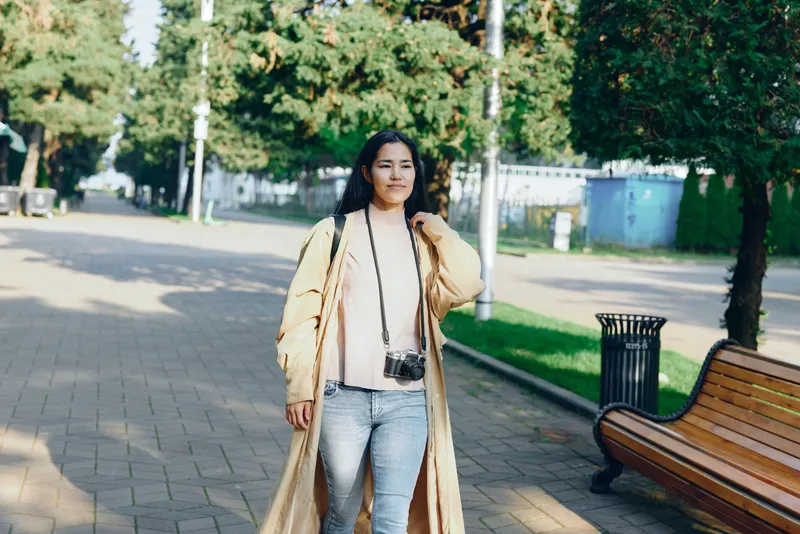 Woman walking in a sunny park wearing a long beige coat and jeans, with a camera hanging from neck, enjoying a casual outdoor stroll along a paved path.