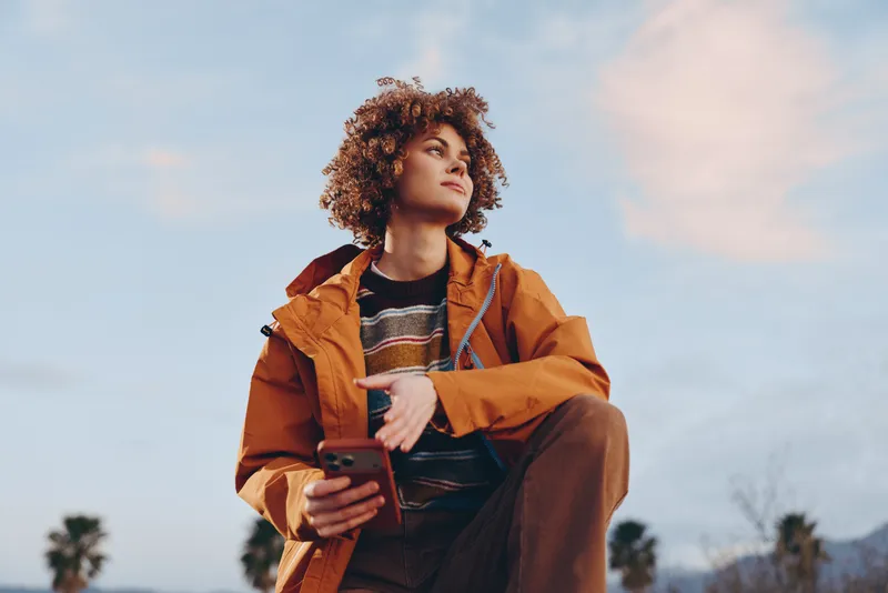 Woman curly hair outdoors, orange jacket striped sweater, lifestyle moment, she holds instrument near body, wide sky background, confident relaxed sunny mood
