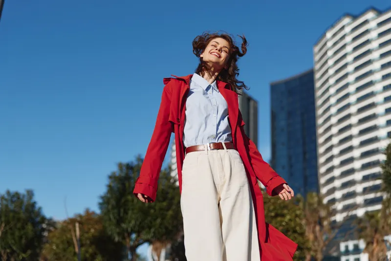 Joyful woman outdoors in a red coat, white pants, blue shirt, smiling under bright sunlight, wind in hair, modern buildings in the background