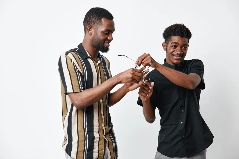 Two young black men with dark skin smiling and interacting while holding eyeglasses on white background. Casual clothing and friendly gesture in studio setting.