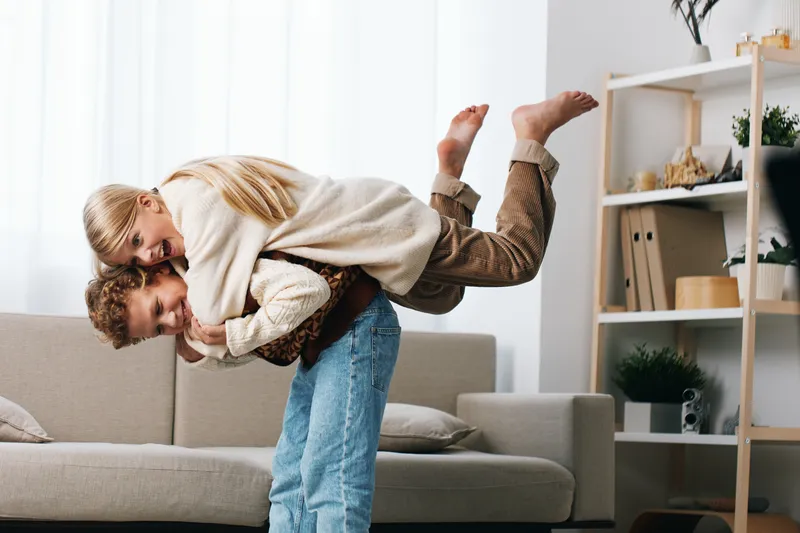 Joyful couple playfully embraces in a cozy living room, the man lifting the partner in a lighthearted moment, soft natural light, warm casual clothes, home vibe.
