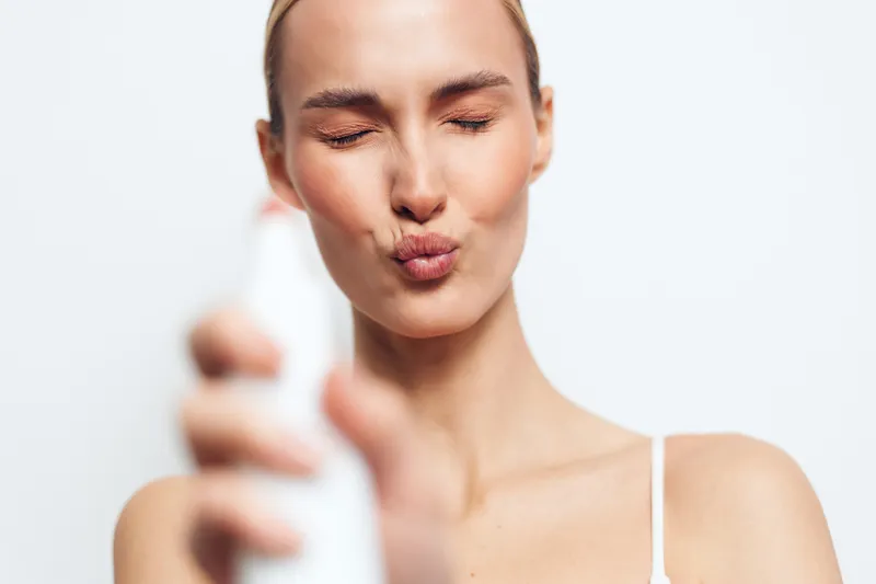 A closeup of a woman with pursed lips spraying a cosmetic bottle, highlighting skincare routine, facial care, and a moment of refreshment in a bright studio setting.