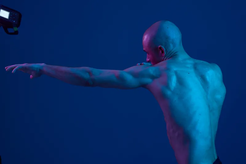 Athletic man posing in dynamic posture, showcasing muscular back against a vibrant blue background with dramatic lighting. A powerful black and white style image.