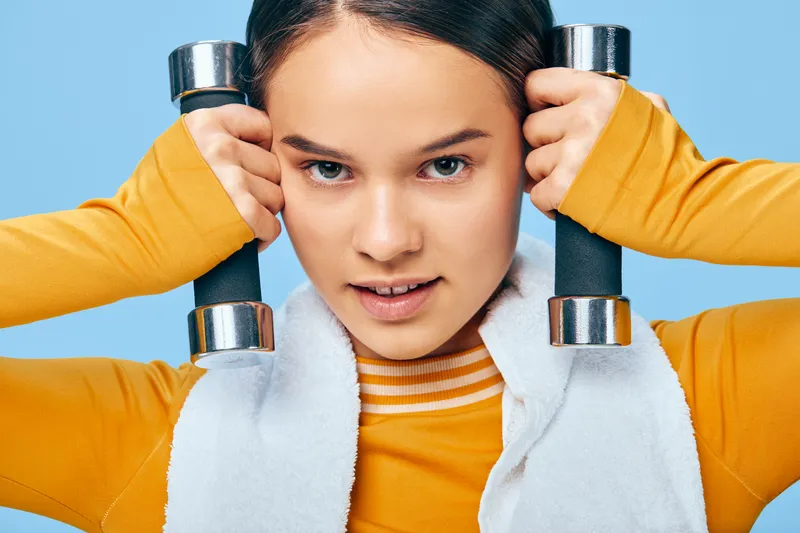 Active woman in bright orange sportswear holds dumbbells near her head, wrapped in a towel, ready for a workout in a bright blue studio, showcasing strength, focus and athletic determination.
