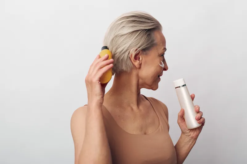 Mature woman with short blonde hair holds two skincare bottles applying cream on face. Concept of beauty routine, facial care, anti aging and personal wellness at home.