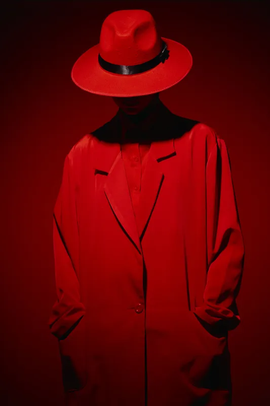 Fashion model in a bold red coat and wide-brimmed hat stands against a red background. The dramatic lighting and strong shadows create a mysterious beauty creative concept.