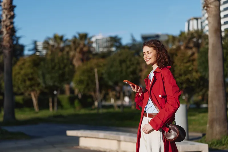 Curly haired woman in a bright red coat smiles while walking in a sunny park, holding a smartphone, conveying casual chic and urban wellness and confidence.
