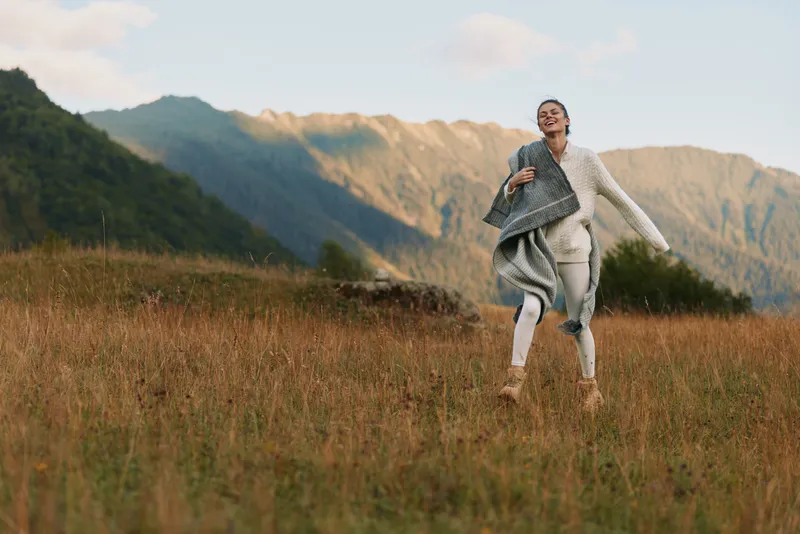 Young woman with long hair joyfully runs through a grassy field in casual clothes during golden hour with mountains in the background and a relaxed expression on her face.
