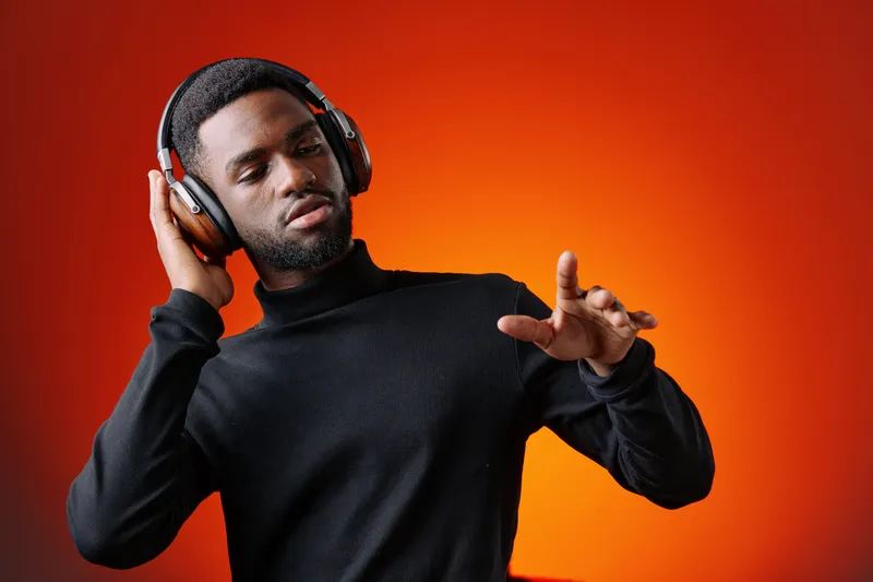 Young Black man enjoying music with headphones against a vibrant orange background, expressing joy and relaxation through his movements and smile