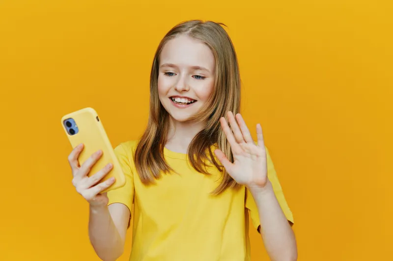 Cheerful girl in yellow shirt smiling and waving while using smartphone against vibrant yellow background, showcasing joy and connectivity in modern communication