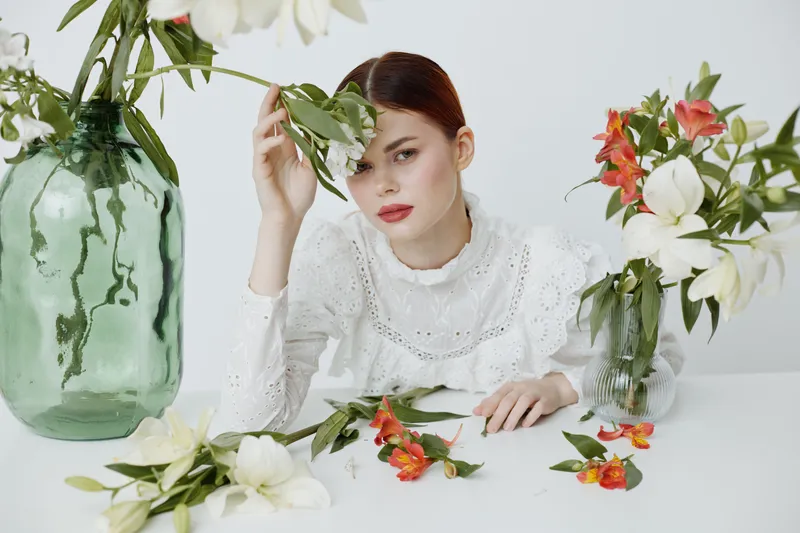Elegant young woman with red hair in a white lace blouse surrounded by floral arrangements, conveying beauty, nature, and femininity in a soft color palette