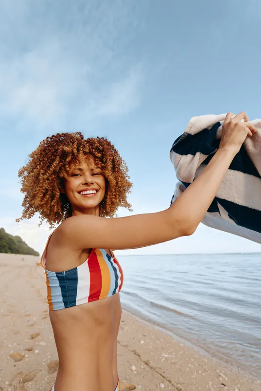 Joyful woman on a sunny beach, wearing a striped bikini, holds up a striped towel, smiling with carefree summer mood, waves wash the shore under a bright blue sky