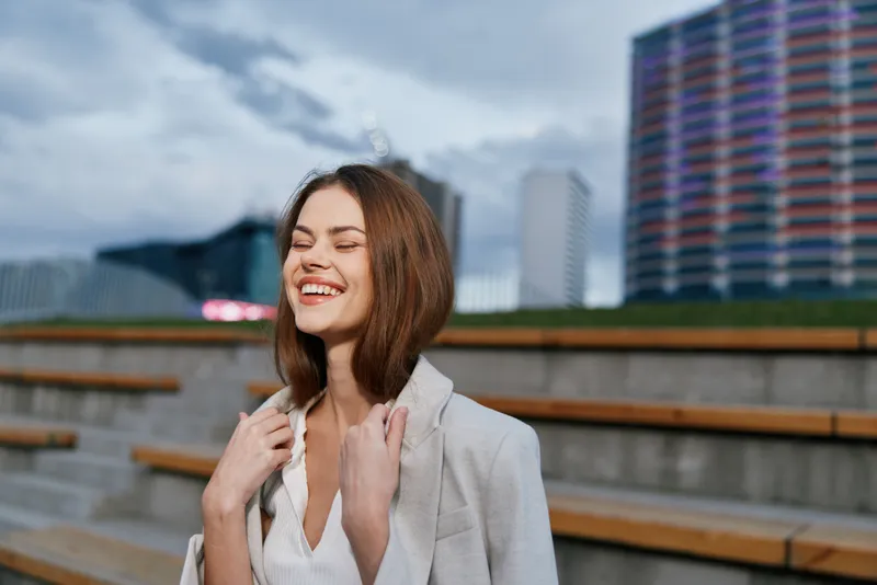Young woman in light blazer smiling outdoors in urban setting with relaxed happiness and natural expression, casual fashion style, spring or autumn season