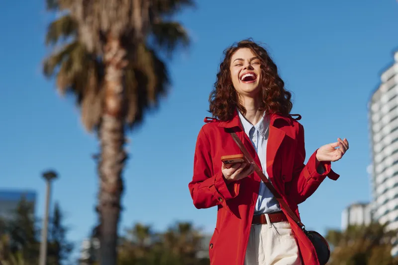 Joyful young woman with curly hair in a red coat laughs while looking at her phone outdoors on a sunny city street, radiating confidence and energy.