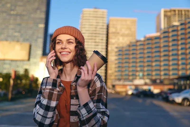 Woman with coffee on phone smiles in urban street during golden hour, candid moment and authenticity captured; warm glow, casual beanie and plaid jacket convey mindful living and joy.
