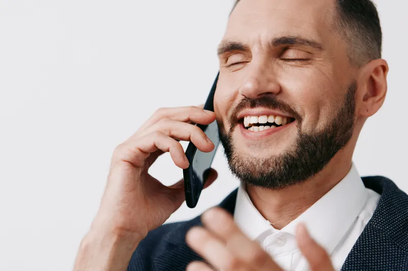 A professional man in a business setting talks on a mobile phone, wearing a smart blazer, and shows a confident smile against a solid color background. The scene conveys success, focus, and friendly