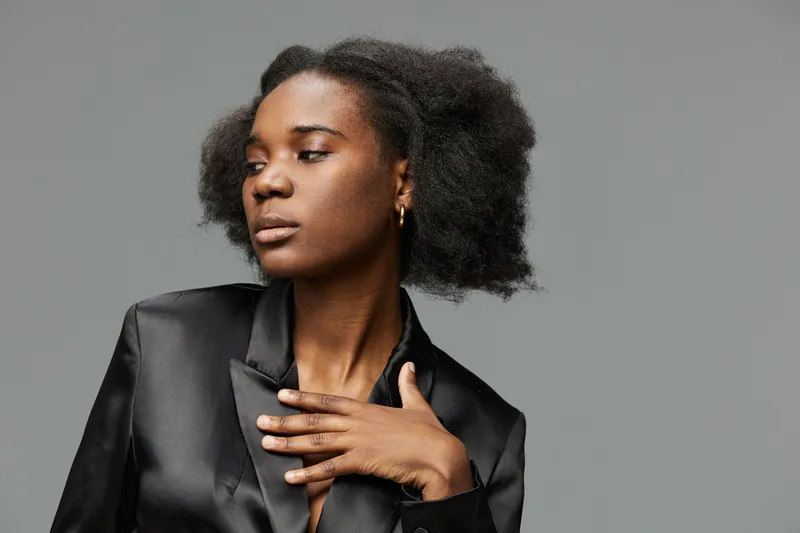 Elegant African woman with natural hair in a black satin jacket, posing gracefully against a minimalistic grey background showcasing confidence and style