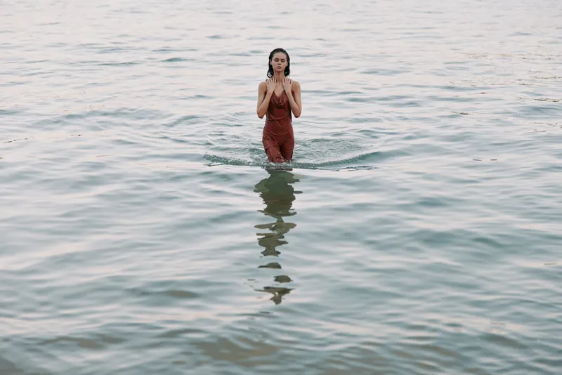 Solitary woman stands in shallow water, red dress billowing softly, hands near chest, tranquil reflections, gentle light, intimate scene of calm solitude.