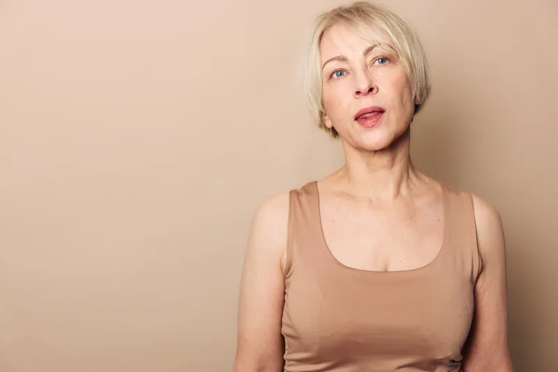 Portrait of a mature blonde woman in a beige tank top against a neutral background. Natural look with minimal makeup, soft lighting highlights calm and confident expression.