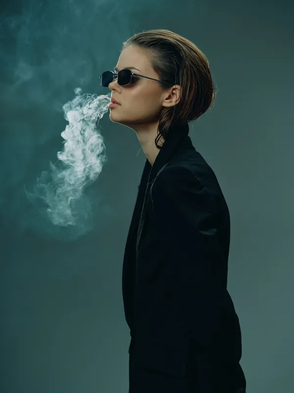 Stylish woman in black smoking with sunglasses and smoke cloud in moody dark background