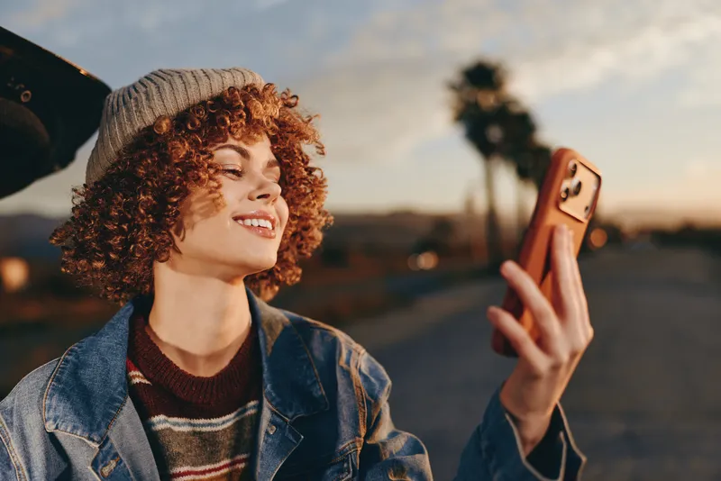 Curly haired woman lifestyle outdoors at sunset smiling while holding a smartphone in her hand denim jacket striped shirt warm golden light open street background relaxed mood city evening