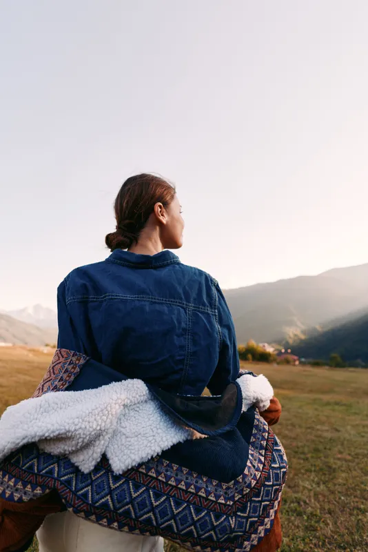 Woman sitting with her back to camera in a denim jacket and patterned blanket, overlooking mountains and sunset landscape in nature, peaceful outdoor travel and solitude.