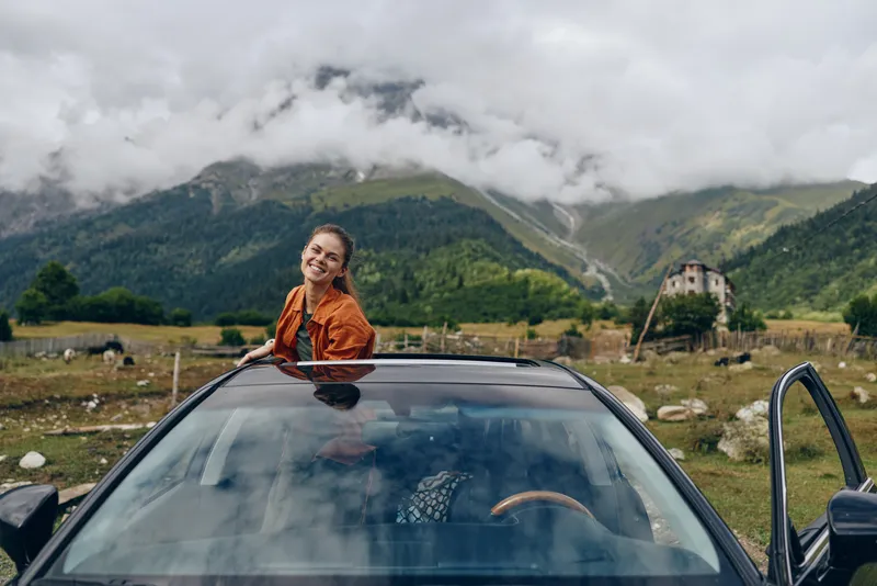 woman car mountains countryside smile sunroof travel roadtrip, joyful young woman standing through car sunroof with open arms in rural valley, authenticity inclusivity, moody clouds and green peaks