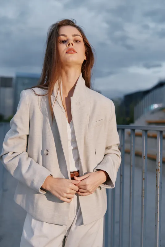 Confident woman in light beige jacket standing outdoors with serious expression during cloudy day urban fashion business style