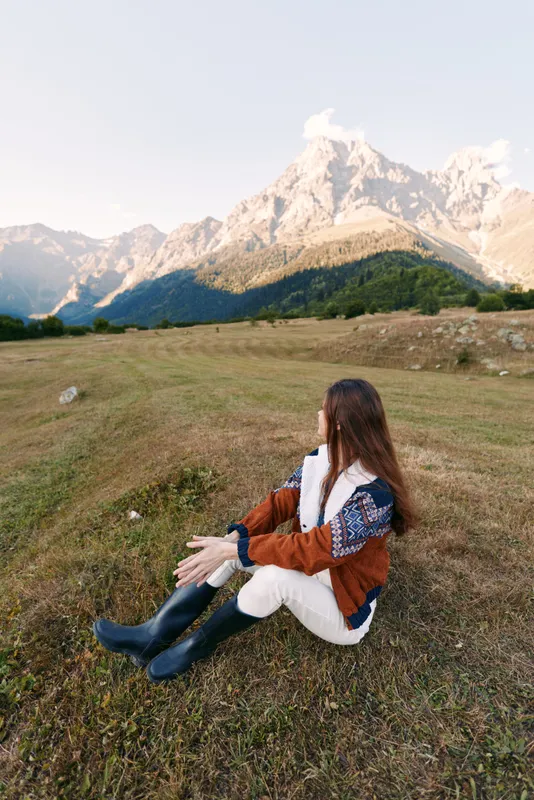 Woman sits on meadow facing distant mountains in a wide landscape of nature. Autumn jacket and rubber boots, solitary traveler enjoying calm outdoor scenery and mountain view.