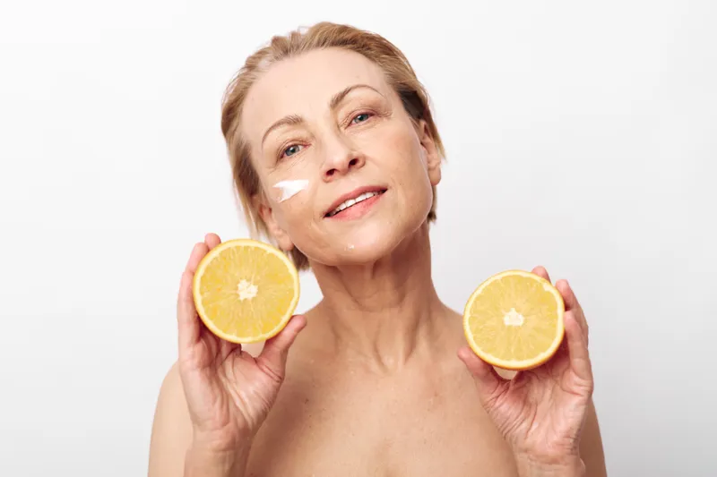 Mature woman with natural skin holds two orange halves near face, applying white cream on cheek, isolated on white background. Concept of skincare, vitamin C, healthy aging, and natural beauty.