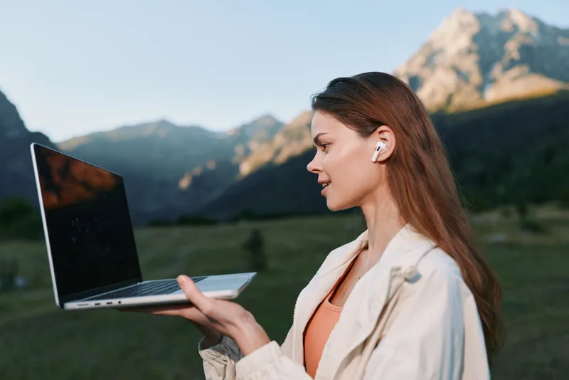 A focused professional woman works outdoors with a laptop in a mountainous landscape. She wears wireless earphones and a light jacket, presenting a modern, productive, and optimistic image of remote