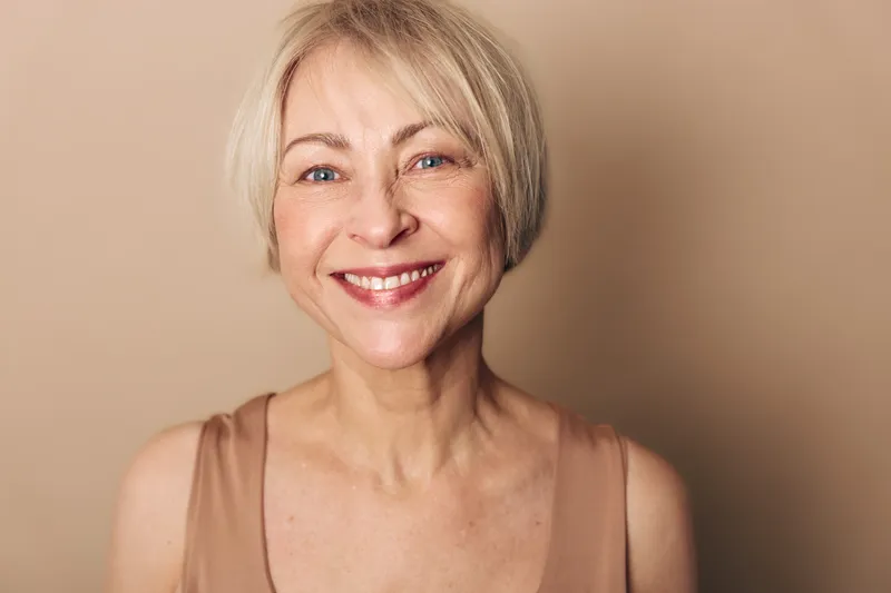 Smiling mature woman with short blonde hair looks directly at camera, natural makeup and warm beige background. Concept of confidence, happiness, beauty, and aging gracefully.