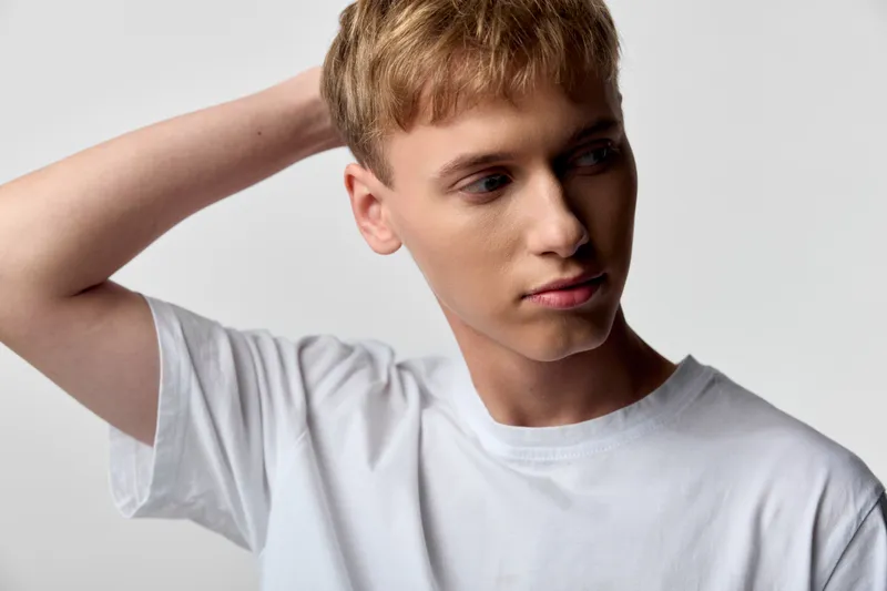 Thinking, casual man with a hand behind the head in a white t-shirt against a neutral studio backdrop, capturing a calm, introspective mood suitable for fashion, lifestyle and creative imagery.