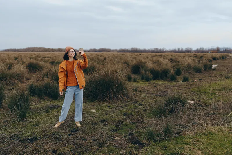 Woman lifestyle outdoors in a wide field with tall grass, wearing an orange jacket and hat, walking along a rural path under soft daylight, natural scenery for casual fashion.